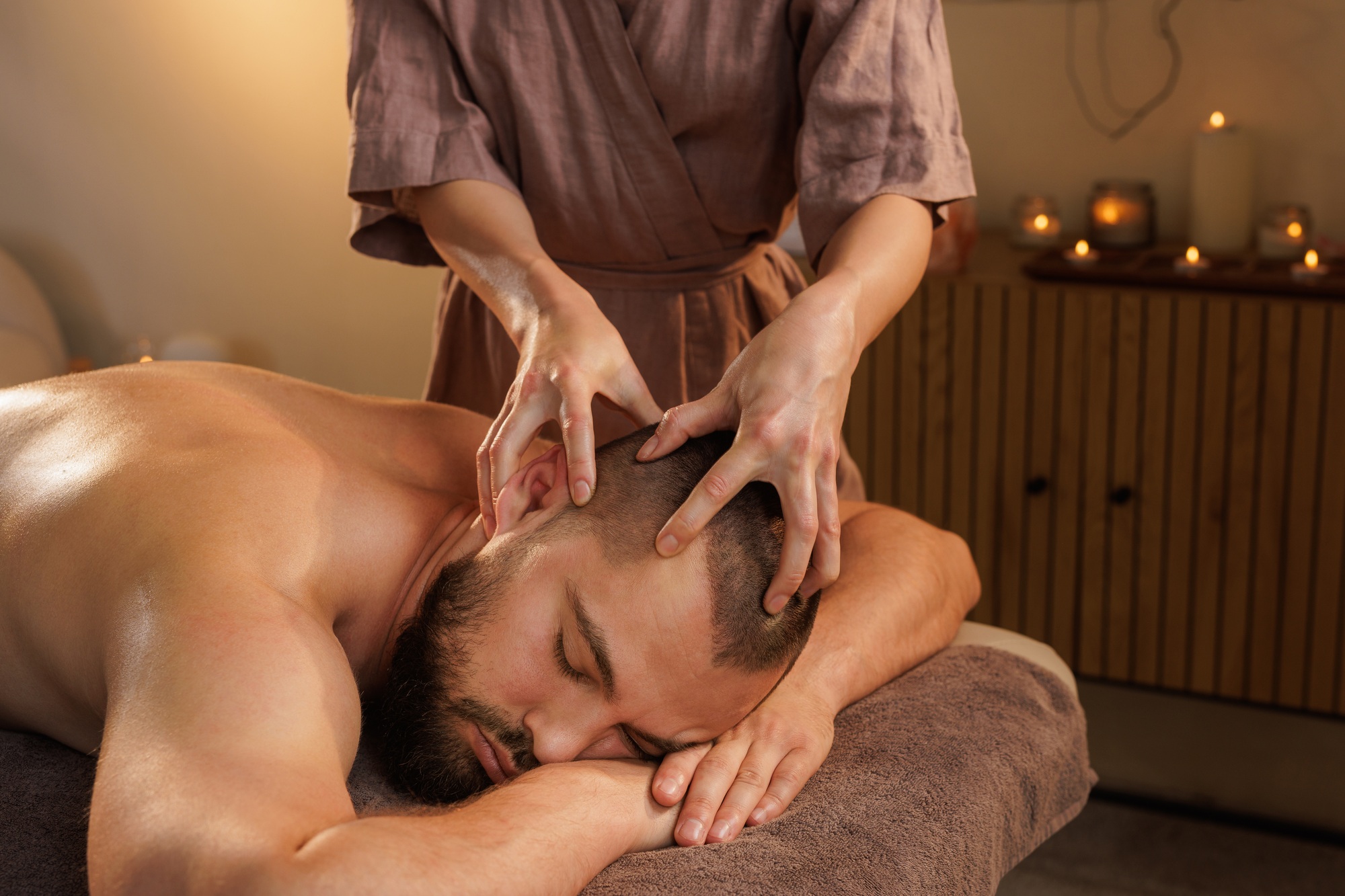 Therapist giving head massage to a man in a calm spa room with candlelight ambiance.